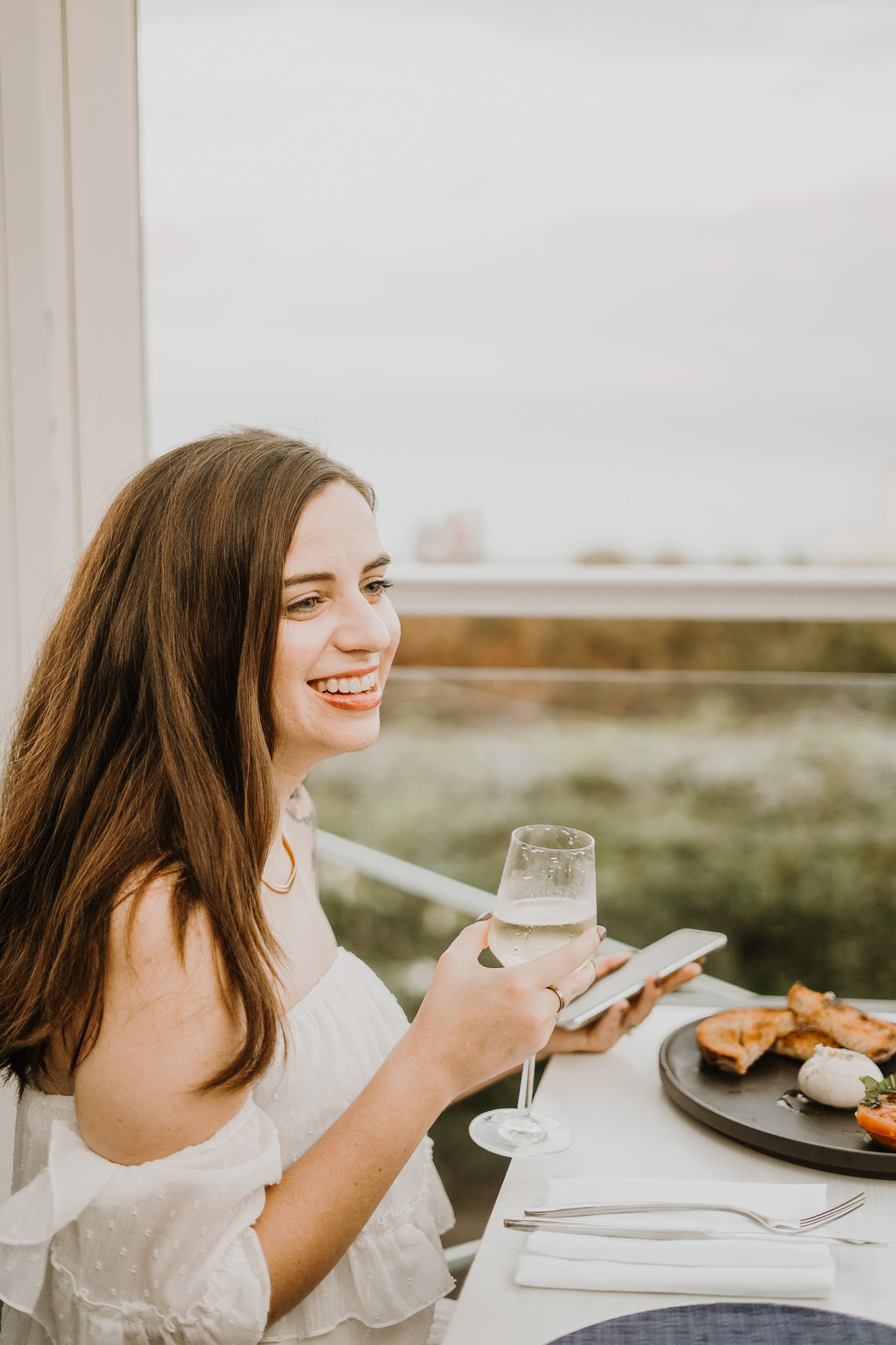 Amanda Burg smiles over a glass of white wine and a plated meal on a sunny terrace, a relaxed scene for a med spa website post