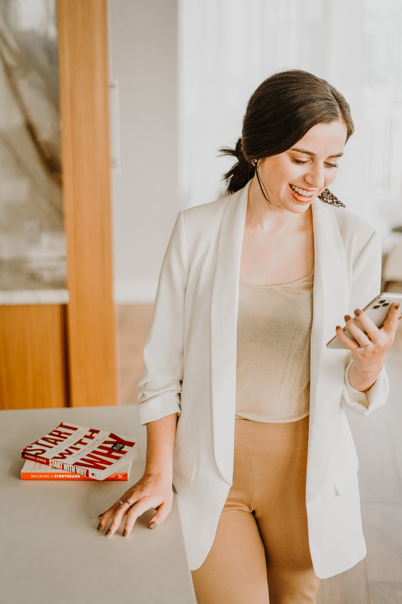 Amanda Burg in a white blazer smiles at her phone beside a stack of books, considering the question: is a rebrand worth it?