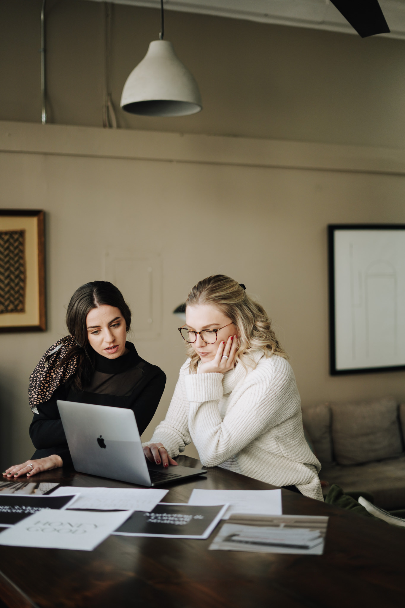 Two women work at a laptop surrounded by printed pages in a warm studio, reviewing ideas to improve a practice homepage